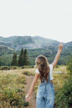 a girl in overalls walking up a dirt path towards the mountains with her arms raised