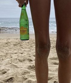 a woman standing on top of a sandy beach next to the ocean holding a green bottle