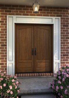 two wooden doors are on the side of a brick building with pink flowers in front