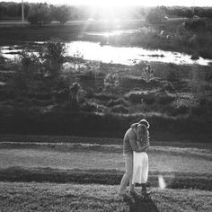 a man and woman are standing in the grass near a river, with sun shining down on them