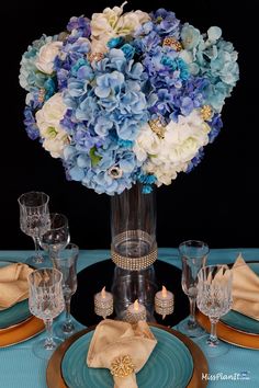 blue and white flowers are in a glass vase on a table set with gold place settings