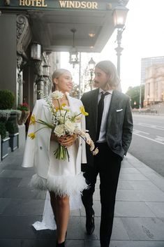 a man and woman are walking down the street holding flowers in front of a hotel