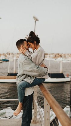 a man and woman hugging on a pier near the water with boats in the background