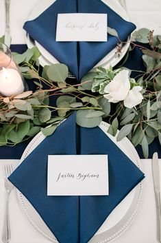 a place setting with blue napkins and greenery on the table for an elegant wedding