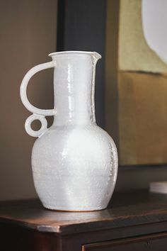 a white vase sitting on top of a wooden table next to a framed art piece