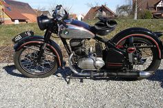 an old motorcycle is parked on the gravel near some grass and houses in the background