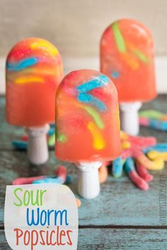 three popsicles covered in colorful candy sitting on top of a wooden table with the words sour worm popsicle