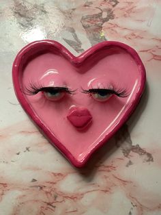 a pink heart shaped bowl with eyes and eyelashes on top of a marble countertop