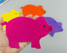 a hand holding three different colored pig magnets on top of a gray table with markers and pencils in the background