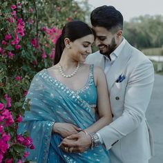 a man and woman standing next to each other in front of pink flowers with their arms around each other