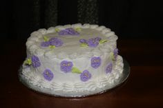 a cake with white frosting and purple flowers on the top is sitting on a table