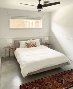 a white bed sitting under a window next to a rug and table with two lamps