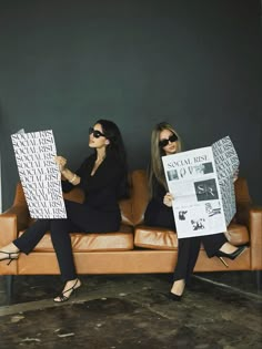 two women sitting on a couch holding up newspapers