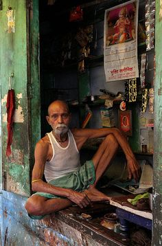 an old man sitting in the doorway of his house with no shoes on it and looking at the camera