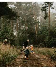 two children running down a dirt road in the woods with an adult standing behind them