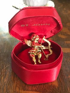 "Here is a very rare example of a St John Cupid brooch from the 1980s. It is in its original red satin box with a good beaded St. John logo on the cover. The Cupid is darling with his little jeweled bow and arrow, the tip of which is black enamel. The bow also has little pearls and rhinestones and the Cupid is wearing a red rhinestone skirt.Inside the box is a little red bean bag pillow on which the Cupid rests. The brooch is signed with the Saint John logo on the reverse and also the logo is pr Vintage Brooch For Valentine's Day Formal, Gold Brooches For Valentine's Day, Gold Brooches For Valentine's Day Formal, Gold Brooches For Formal Valentine's Day, Antique Red Brooch As Gift, Antique Red Brooches As Gift, Antique Red Brooches As A Gift, Antique Red Brooches For Gift, Red Bean Bag