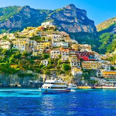 a boat is in the water next to some buildings on a mountain side with mountains in the background