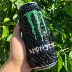 a hand holding up a black and green monster energy drink in front of some bushes