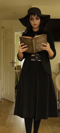 a woman in a black dress and hat is holding an open book while standing on a hard wood floor