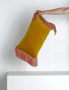 a hand is holding a yellow pillow with fringes on the bottom and an orange one in the middle