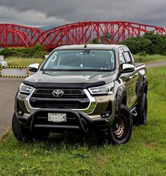 the front end of a silver toyota pickup truck parked on grass near a red bridge