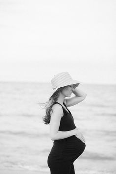 a pregnant woman standing on the beach wearing a hat