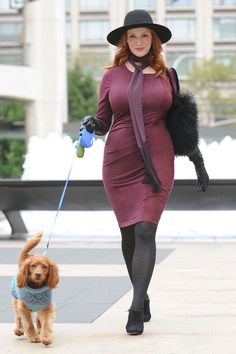 a woman in a dress and hat walks her dog on a leash through the city