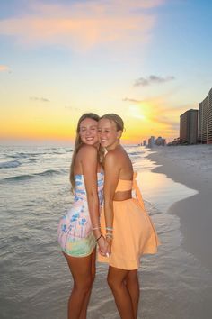 two girls standing on the beach at sunset with their arms around each other's shoulders