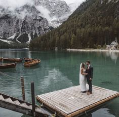 a bride and groom standing on a dock in the middle of a lake surrounded by mountains