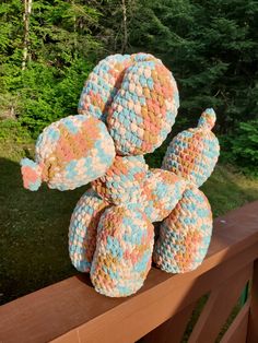 a crocheted teddy bear sitting on top of a wooden rail next to trees