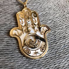 "Hamsa in 24 carat gold on 925 silver and natural white mother of pearl backdrop. In the symbol it is shown Fatima's palm-shaped whose meaning is \"five\". Also named Hand of Miriam, Hamsa or Khamsa, is a very ancient hamulet we can find in many times and different traditions throughout history, predominantly Muslims and Jews, or Cristians but earlier among Egyptians, Sumerian and Babylonian. Its shapes hidden different faces of the feminine cult: as Mother Earth, in the meaning of fertility and Brass Jewelry With Shiny Finish For Gift, Shiny Brass Jewelry Gift, Handmade Symbolic Gold Jewelry, Symbolic White Jewelry With Polished Finish, White Symbolic Jewelry With Polished Finish, Handmade Yellow Gold Mother Of Pearl Jewelry, Luxury Handmade Gold-tone Jewelry, Engraved Mother Of Pearl Jewelry For Gift, Engraved Mother Of Pearl Jewelry Gift