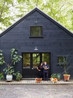 a man and woman standing in front of a black house with two dogs on the porch