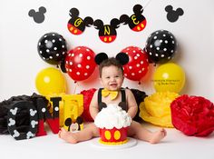 a baby is sitting in front of mickey mouse cake and balloons for his first birthday