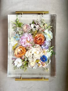 an arrangement of flowers in a square glass box with gold handles on a gray surface