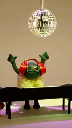 a green monster sitting on top of a bench next to a disco ball hanging from the ceiling