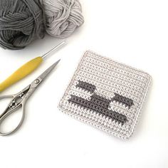 a crochet square next to a pair of scissors and ball of yarn with eyes drawn on it