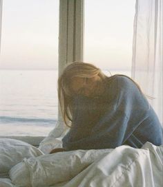 a woman laying in bed with her head on the pillow looking out at the ocean