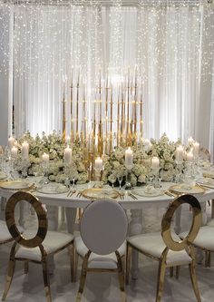 a table with candles and flowers on it in front of a curtained window, surrounded by white chairs