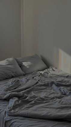 an unmade bed with grey sheets and pillows in a room that has white walls