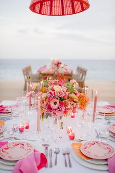 the table is set with pink and orange flowers in vases, candles, and plates