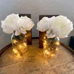 two vases with white flowers are sitting on a wooden table and one has lights in it