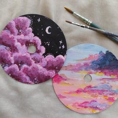 two painted discs sitting next to each other on a white surface with paintbrushes