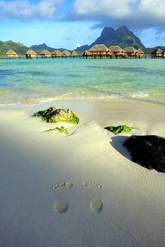 two footprints in the sand next to water and huts with thatched roof overhangs