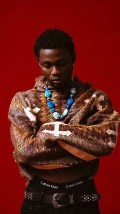 a man with his arms crossed standing in front of a red background wearing a sweater and beaded necklace