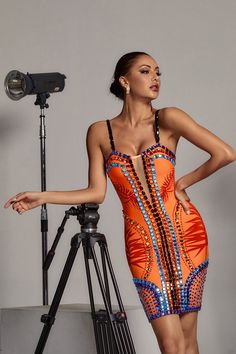 a woman in an orange and blue dress standing next to a tripod with her hands on her hips