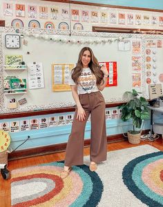 a woman standing in front of a wall with pictures on it and wearing wide legged brown pants