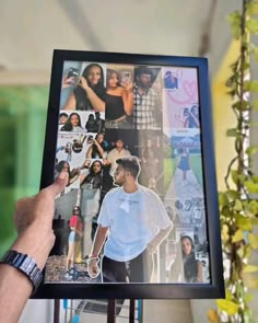 a person holding up a framed photo with many pictures on it and plants in the background