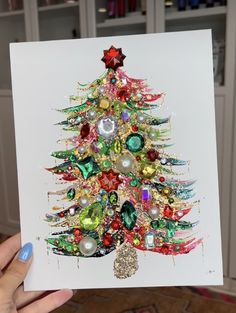 a hand holding up a christmas card with a colorful tree on it's side