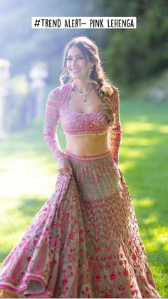 a woman in a pink and gold lehenga standing on the grass with her arms behind her back