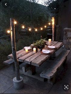 a wooden table with candles and plates on it in front of some lights hanging from the trees